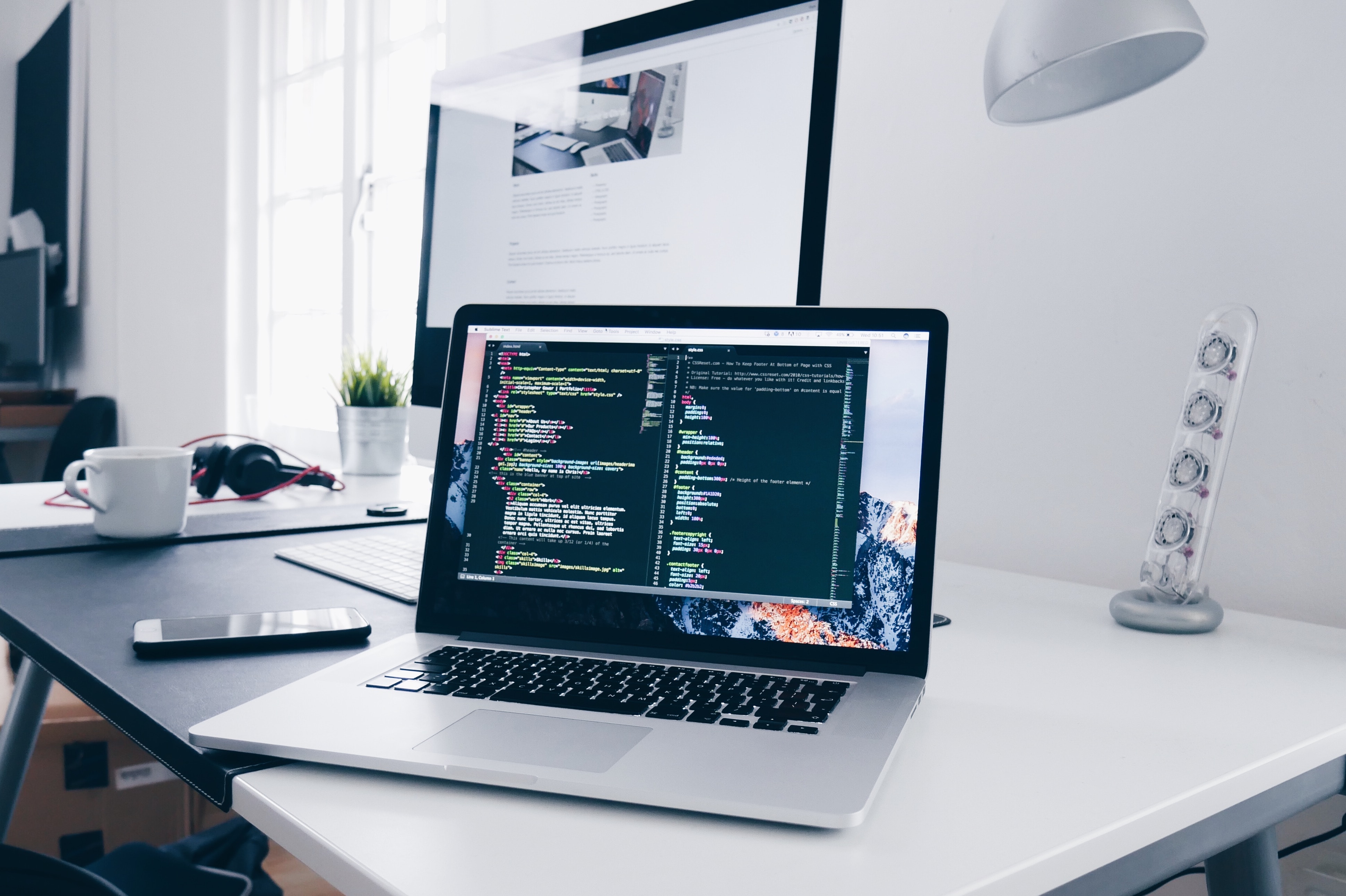 A picture of a person holding a pencil while working on a laptop with code displayed on the screen. They are sitting at a wooden desk with a white wall behind them.